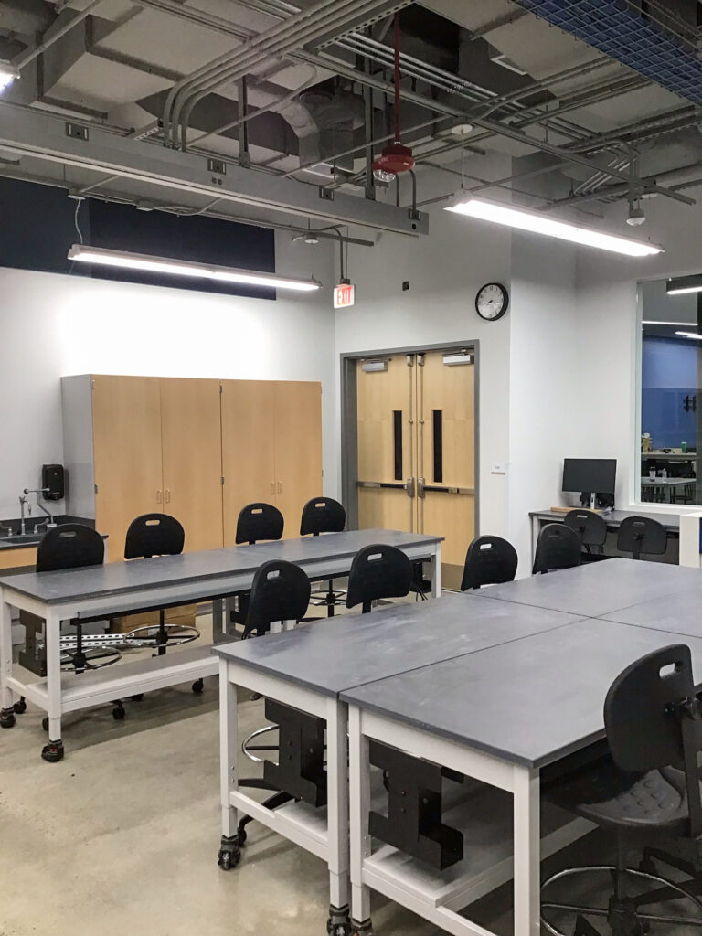 A science classroom with two long grey tables, rolling black chairs, light wood cabinets, and double doors under exposed ceiling pipes.