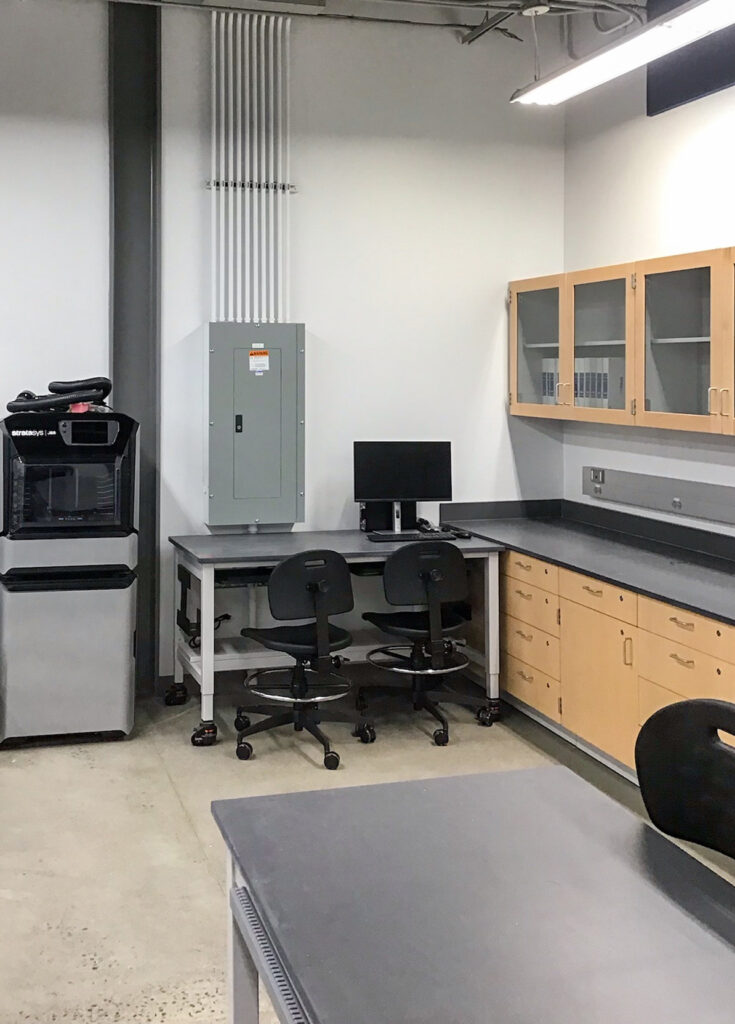 A laboratory workspace with a 3D printer, a computer workstation with two rolling chairs, wall-mounted storage cabinets, and base cabinets.