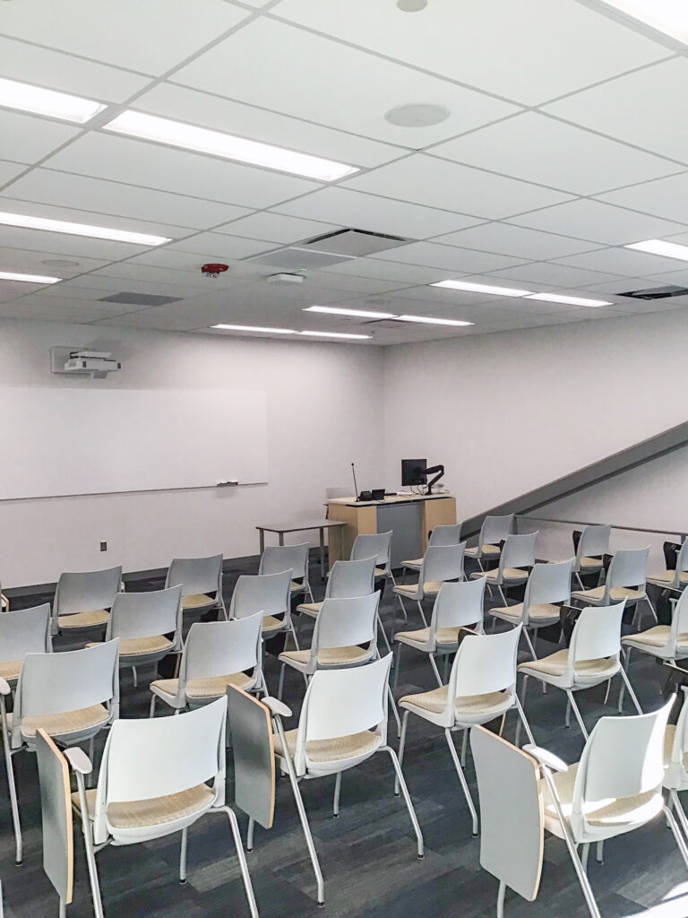 A classroom with rows of white chairs facing a lectern and a whiteboard.