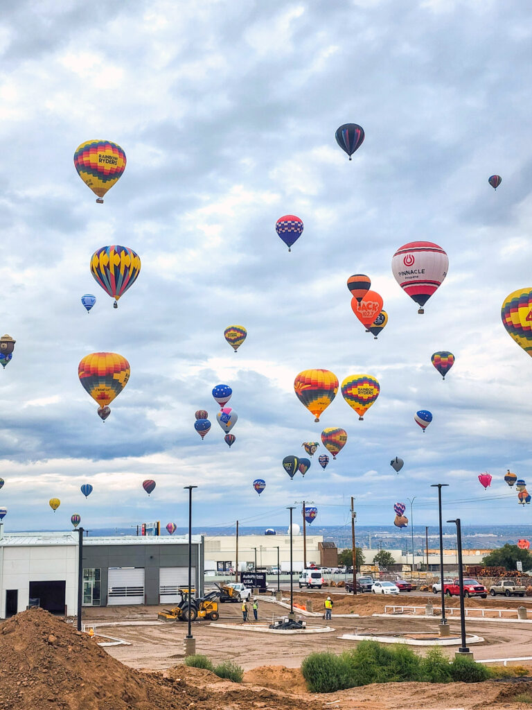 Many hot air balloons floating in the sky above a construction site.