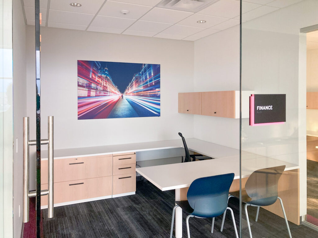 A small office with a white desk, wood storage cabinets, two chairs, and a large wall print of city light trails. A sign on the glass door says “FINANCE.”