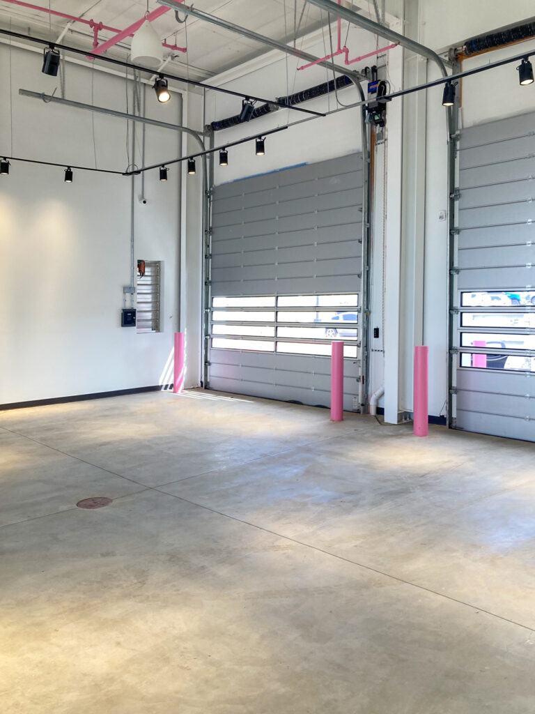 An empty commercial space with concrete floors, white walls, exposed ceiling pipes painted pink, and two large gray roll-up doors.