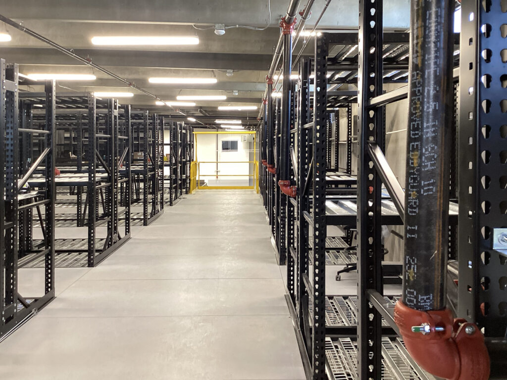 Rows of empty industrial metal shelving units in a warehouse with a yellow safety gate at the end of the aisle.