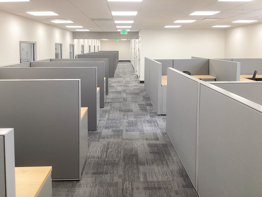 Rows of gray office cubicles with light wood desks in an open-plan office.