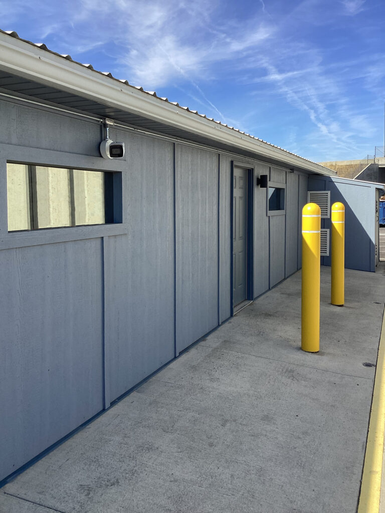 Blue shed exterior with a security camera, a door, and two yellow bollards on a concrete walkway.