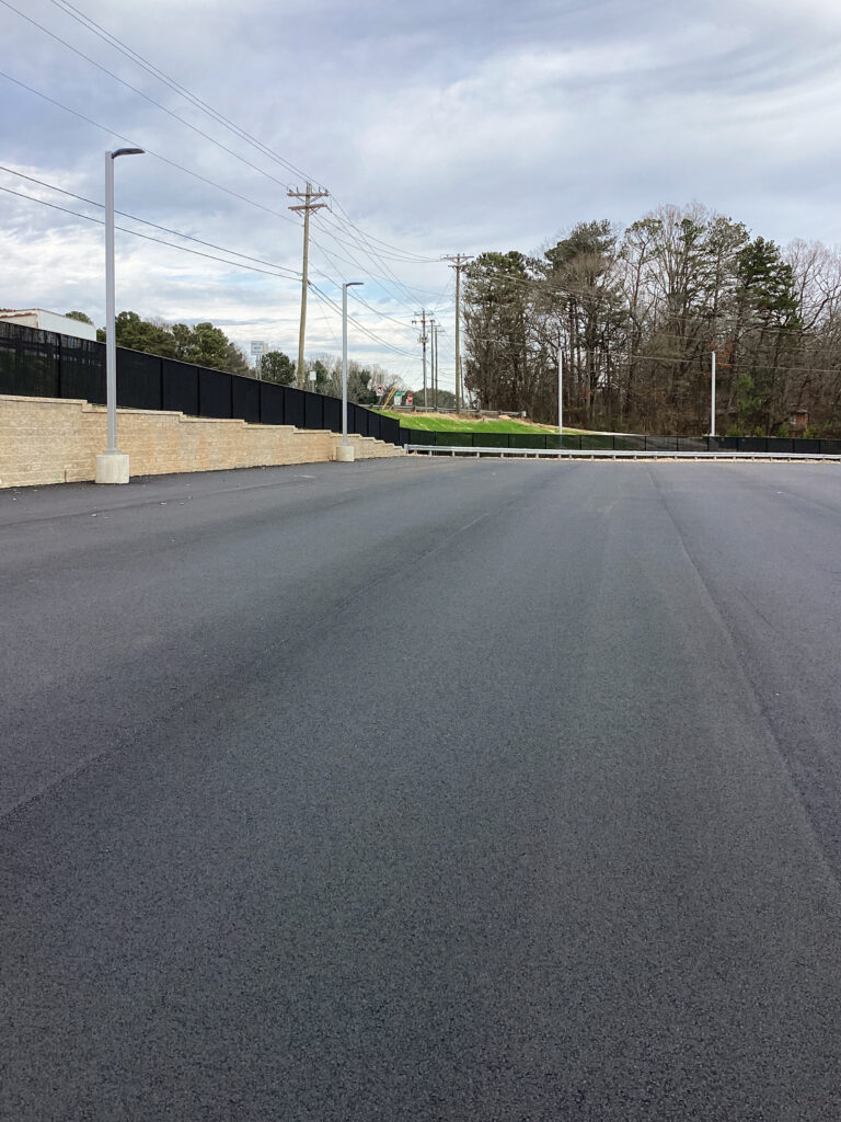 A freshly paved asphalt parking lot bordered by a low stone retaining wall, light poles, and a black fence.