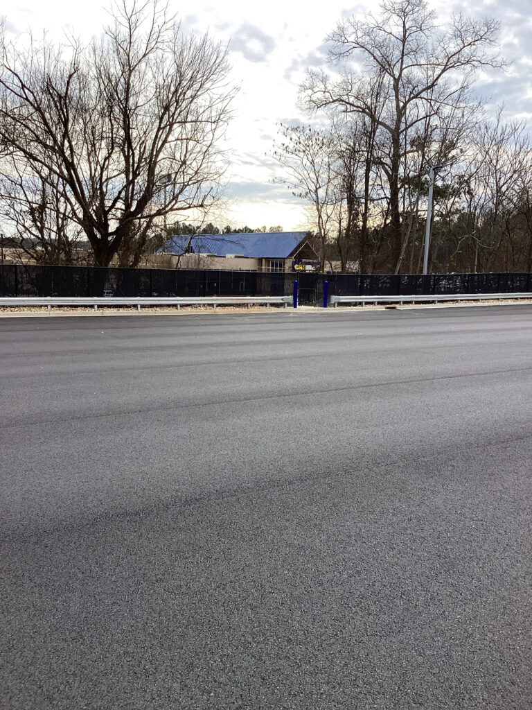 A view of a building with a blue roof, seen from across a wide, freshly paved asphalt road and a metal guardrail.