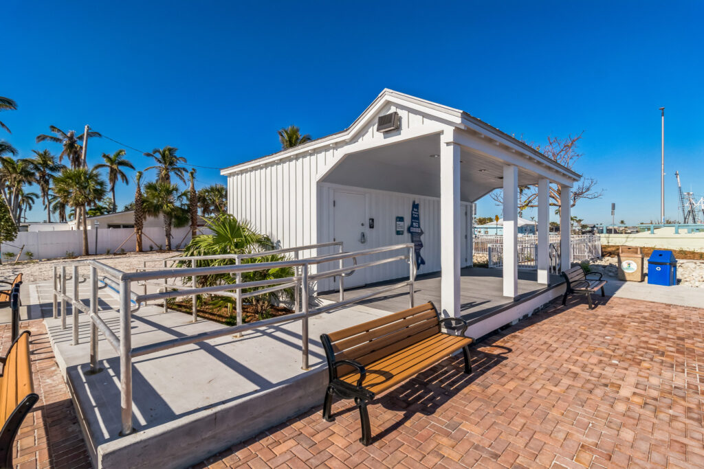 A white park building with a wheelchair ramp, a covered porch, and wooden benches on a brick plaza.
