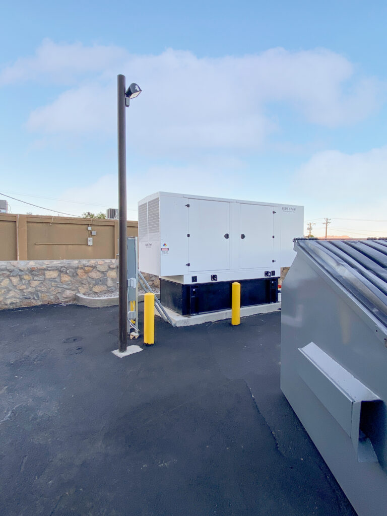 White industrial backup generator installed on a concrete pad between a stone wall and a large gray dumpster.