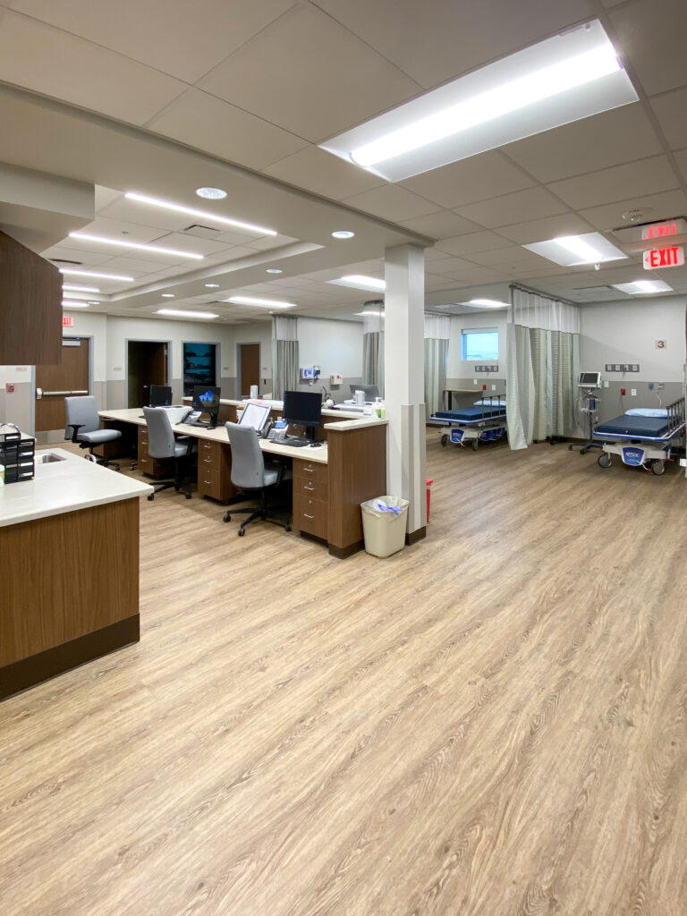 A medical workspace with a central nursing station containing several computer desks, flanked by hospital patient bays with beds separated by privacy curtains.
