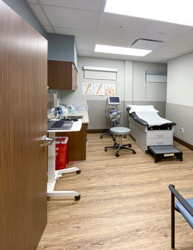 An examination room with an exam table, a rolling stool, medical equipment, and a computer on a mobile workstation.