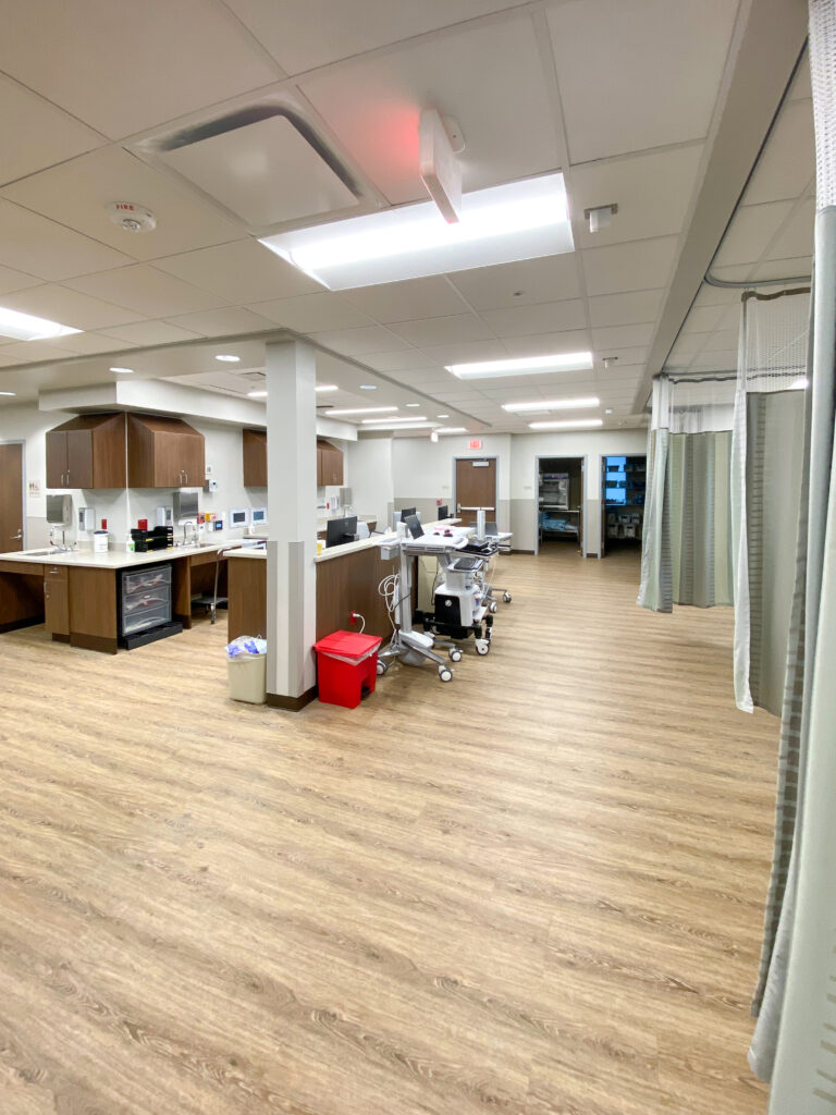 A clinical workspace with wood-look flooring, counters, and medical equipment on wheels, including a red sharps container.