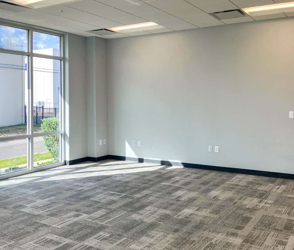 Empty office room with grey walls, patterned carpet, and large windows.