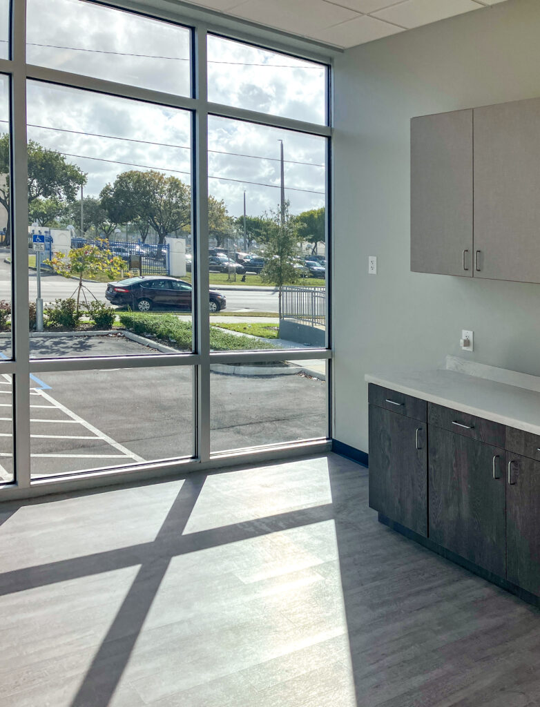 A room with a large window overlooking a parking lot, featuring gray lower cabinets and a wall-mounted cabinet.