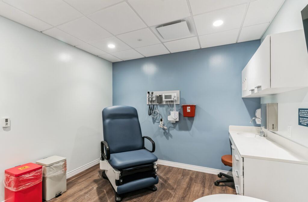 A medical examination room with a blue exam chair, wall-mounted medical equipment, a sink, and a waste bin.