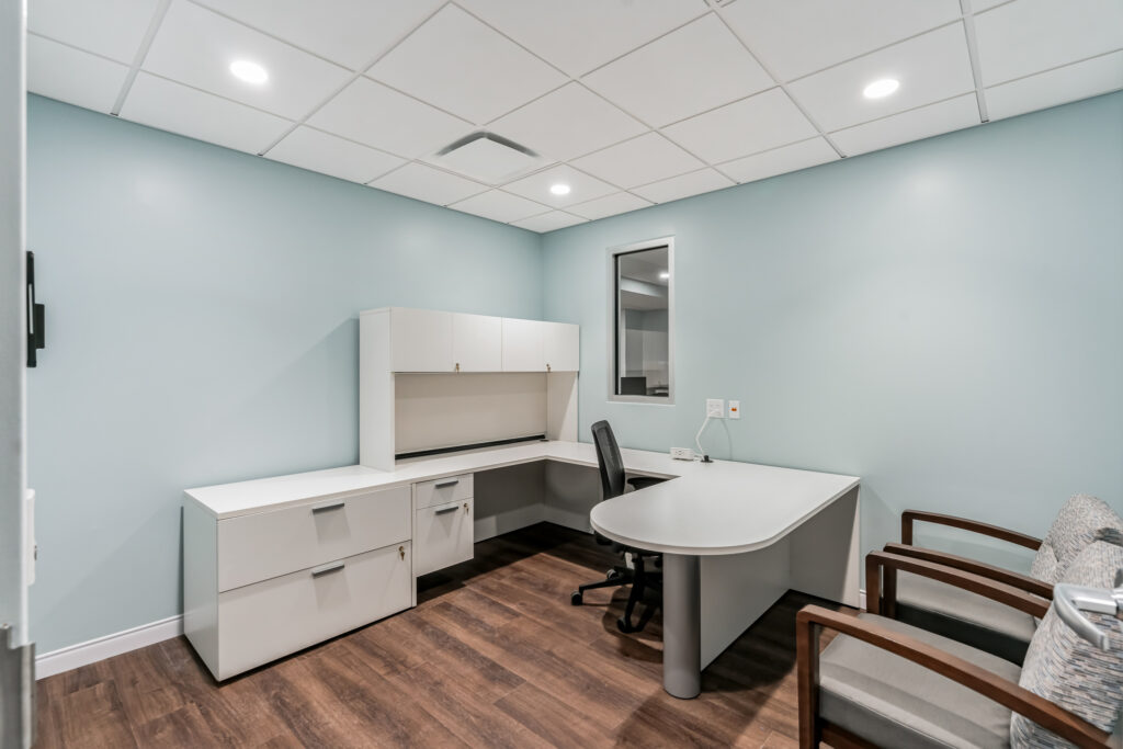 An office with a white L-shaped desk, a black office chair, and two grey armchairs against light blue walls.