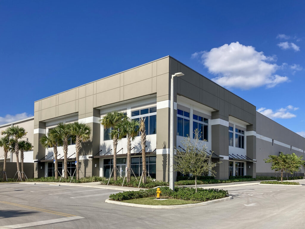 A modern gray commercial warehouse building with large windows and a row of palm trees in front under a blue sky.