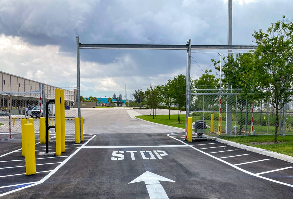 An entrance to a facility featuring a stop sign painted on the asphalt, a directional arrow, yellow bollards, and a metal gate structure under a cloudy sky.