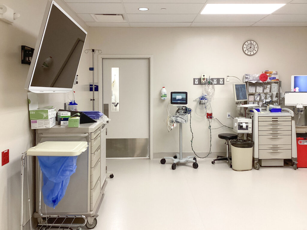 A sterile hospital room containing a medical cart, a patient monitor on a rolling stand, a supply cabinet, and a closed door.