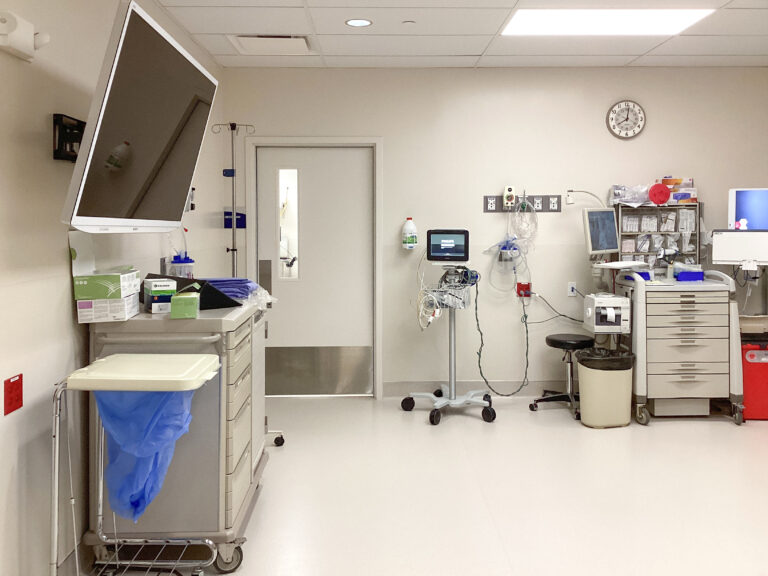 A sterile hospital room containing a medical cart, a patient monitor on a rolling stand, a supply cabinet, and a closed door.