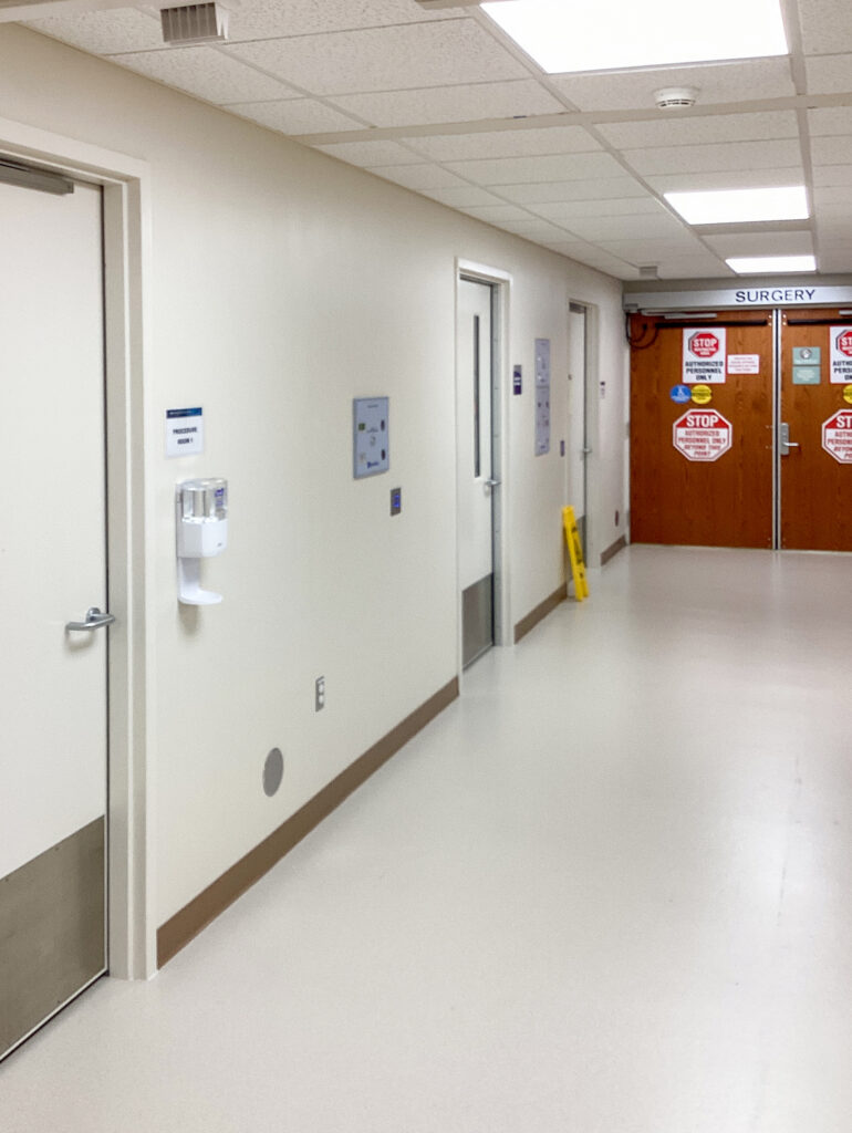 Hospital corridor featuring a hand sanitizer dispenser, closed office doors, and double doors labeled “SURGERY” with red stop signs for authorized personnel only.