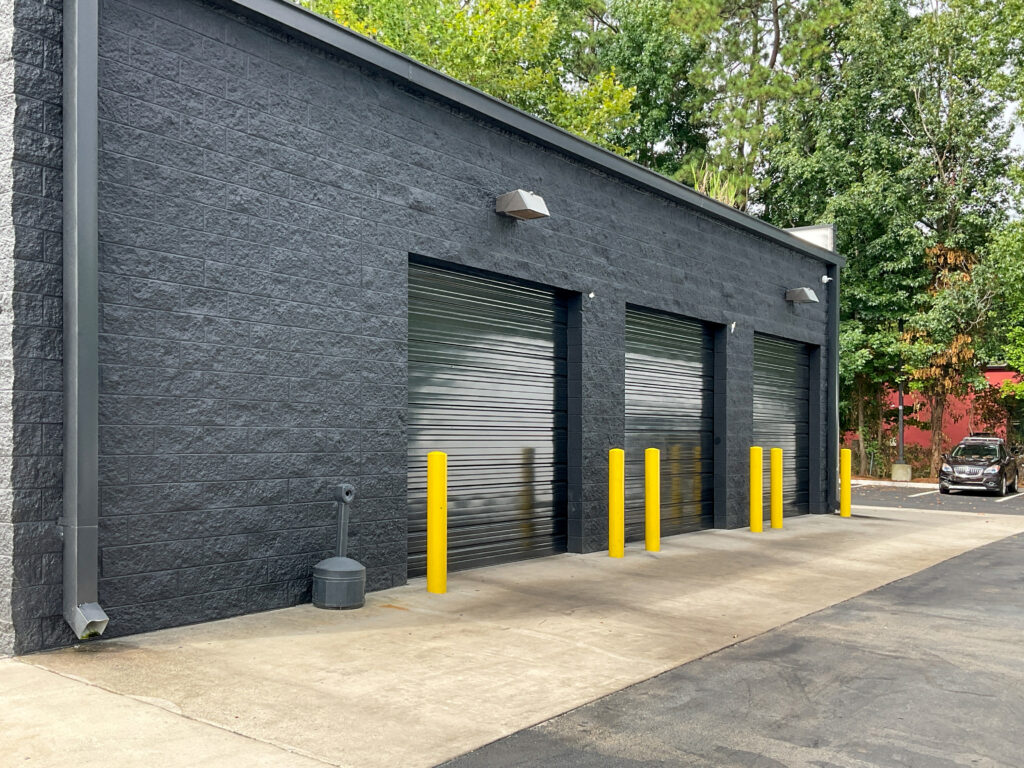 A black building exterior with three closed roll-up garage doors, fronted by yellow bollards on a concrete apron.