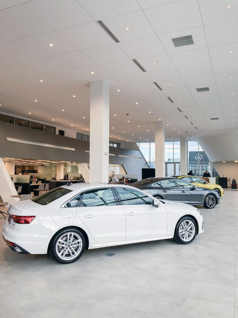 A white sedan, a grey sedan, and a yellow sports car parked inside a brightly lit car dealership showroom.