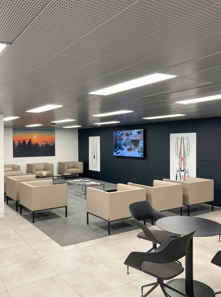 Waiting area with tan armchairs, a glass coffee table, and a television mounted on a black wall with decorative prints.