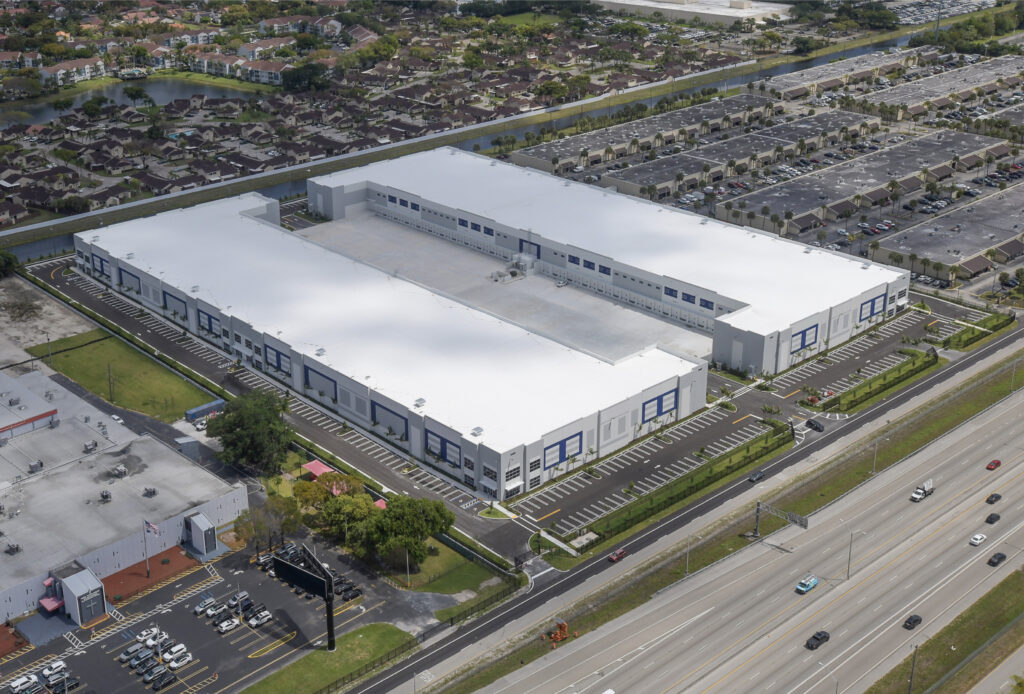 Aerial view of a large white warehouse facility with a flat roof and surrounding parking lots, situated next to a highway.