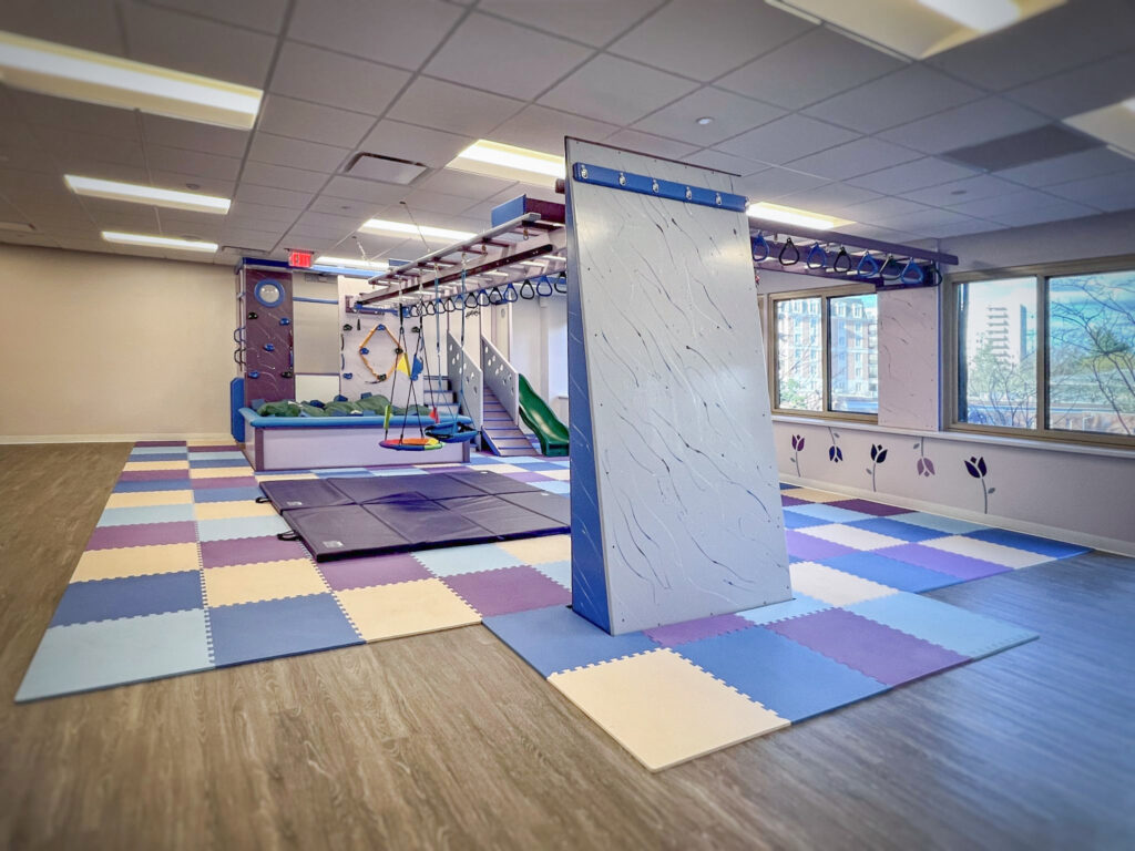 Indoor sensory gym with a rock climbing wall, monkey bars, a hanging swing, a slide, and foam floor mats.