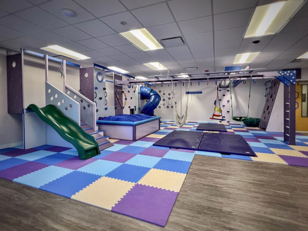 An indoor sensory play gym with a slide, a ball pit, a climbing wall, hanging swings, and a purple and blue foam tile floor.