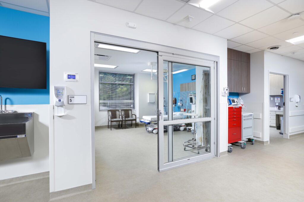 View into a hospital exam room through an open sliding glass door, with a patient bed and chairs visible inside.