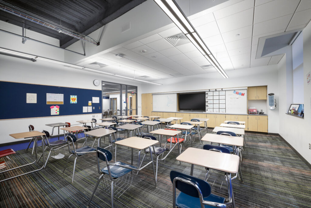 Empty classroom with rows of individual student desks and chairs, a large display screen, whiteboards, and a blue bulletin board.