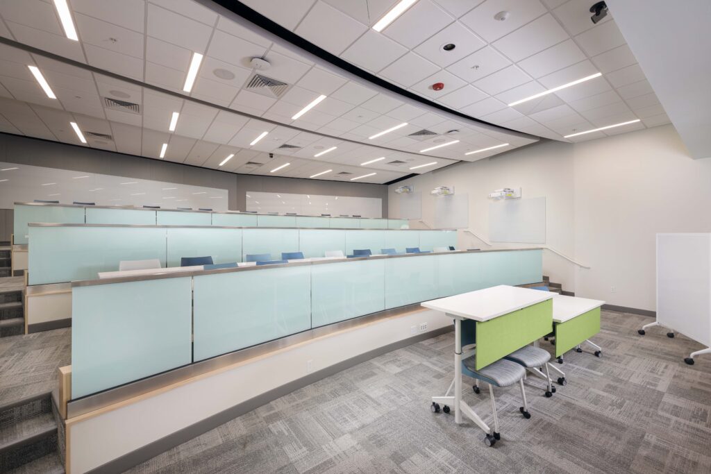 A tiered university lecture hall with rows of pale blue partitions, desk surfaces, and rolling chairs.
