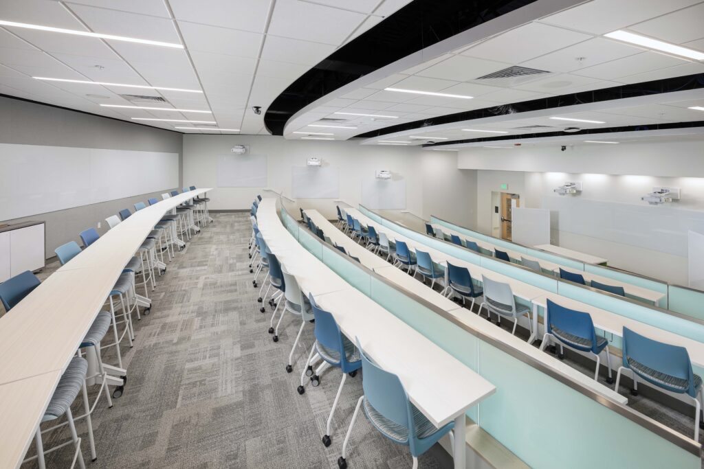 Tiered lecture hall with rows of desks and blue chairs facing whiteboards at the front.