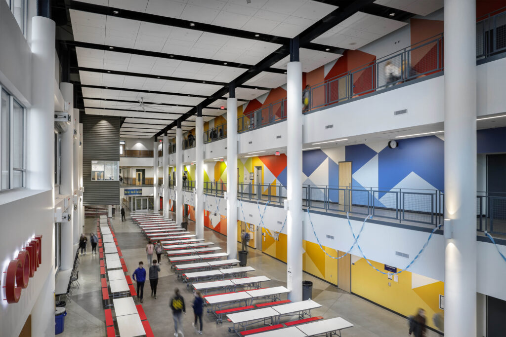 High-ceilinged school atrium with rows of cafeteria tables and colorful geometric wall murals.