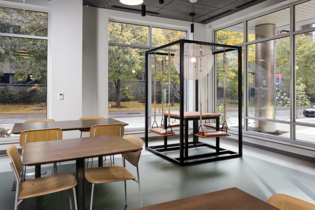 Modern office area with tables, chairs, and a cube-shaped metal frame structure featuring four suspended swing seats around a central table.