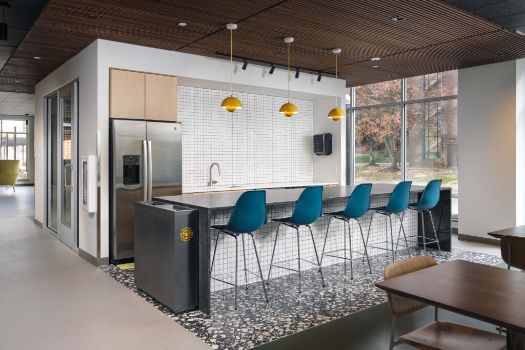 A modern office kitchen featuring a long counter with five blue bar stools, three yellow pendant lights, and a stainless steel refrigerator.