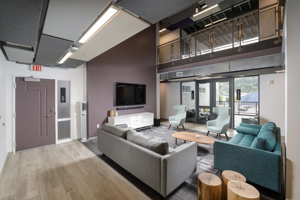 A modern office lounge with a grey sofa, teal sofa, two armchairs, a coffee table, and a wall-mounted television.