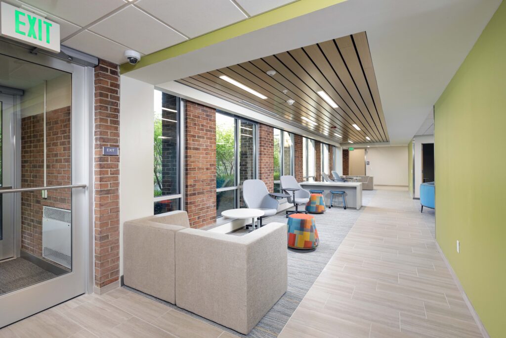 A modern office hallway with brick pillars, windows, seating areas featuring upholstered armchairs and colorful stools, and a light wood-look floor.
