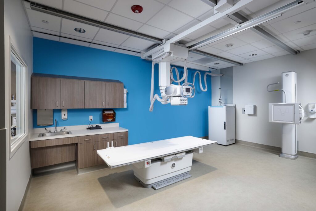 An X-ray examination room with a medical imaging table, an overhead X-ray tube, and a wall-mounted sensor.