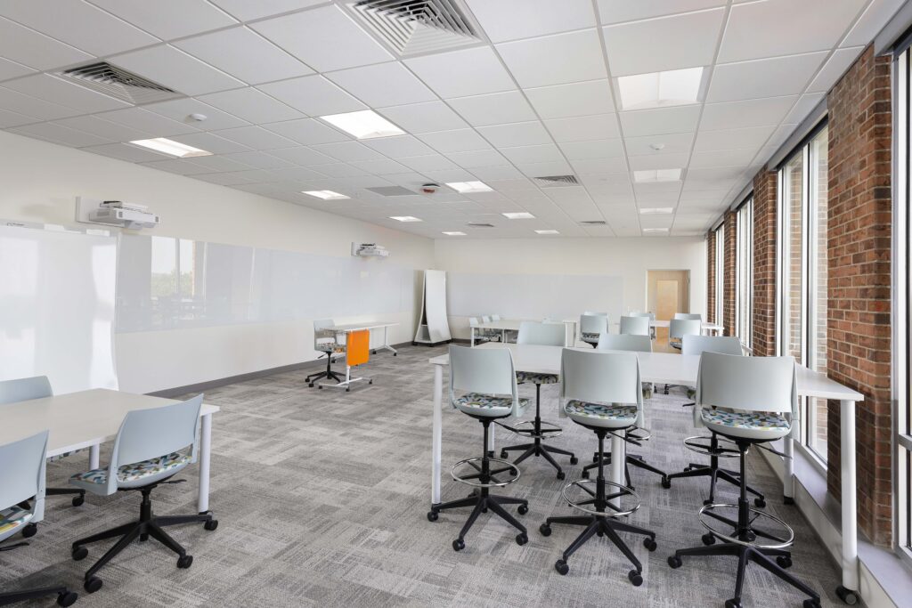 A bright classroom with gray carpet, white tables, and chairs featuring patterned cushions and orange accents.