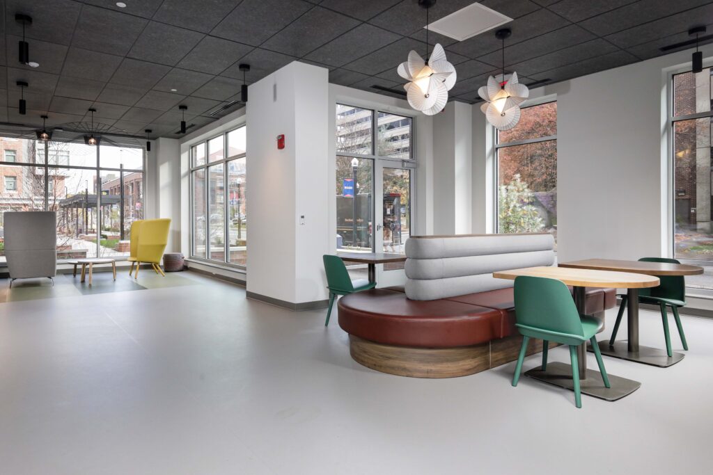 Modern office lobby featuring a curved banquette seating area with green chairs and tables, and a lounge area with yellow chairs near floor-to-ceiling windows.