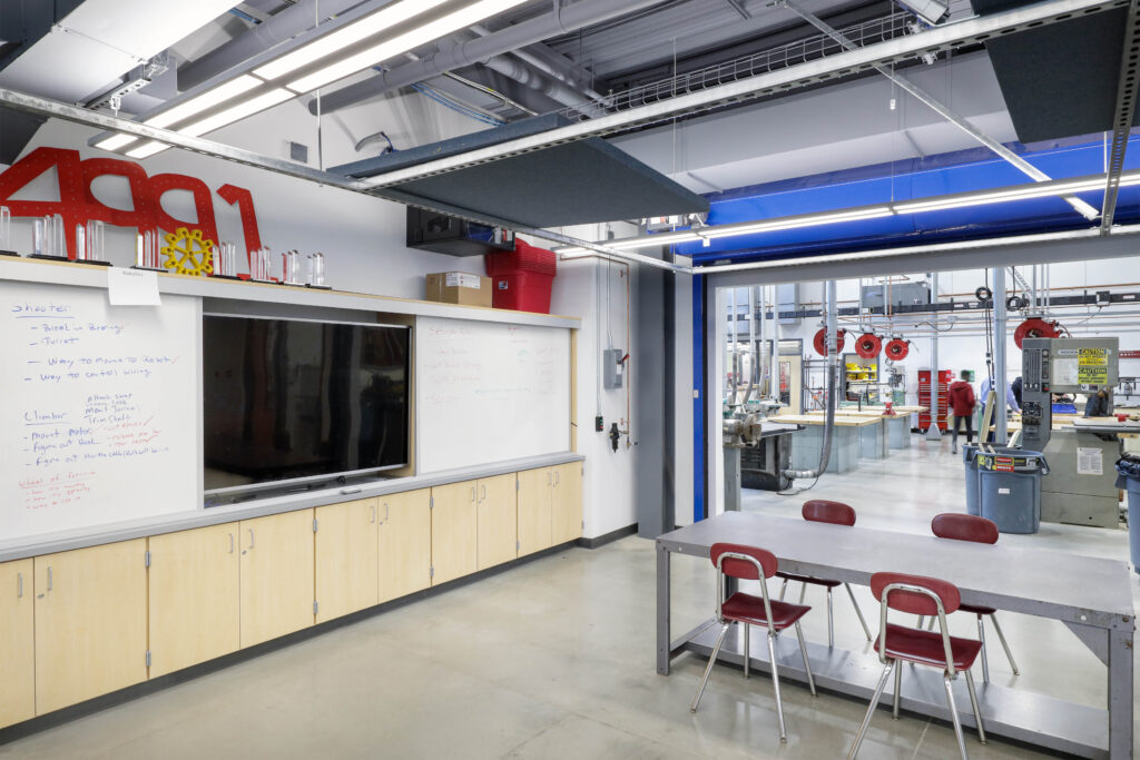 Robotics workshop with a whiteboard displaying handwritten notes, a flat-screen TV, wooden cabinets, and a gray metal table with four red chairs, looking into a machine shop.