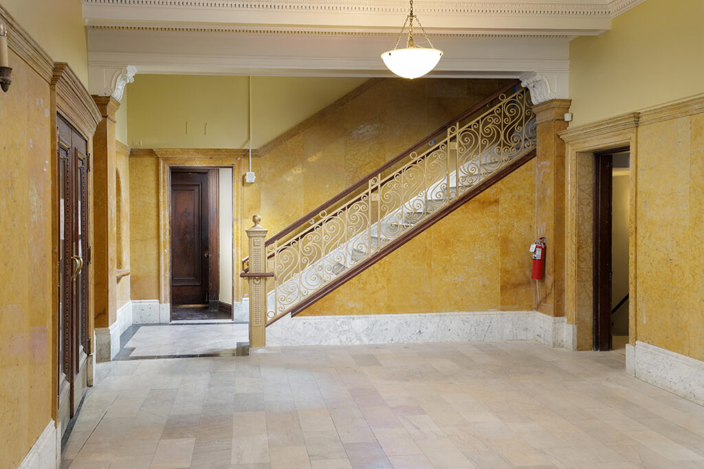 Interior lobby with yellow walls, a stone floor, and a staircase featuring an ornate metal railing.