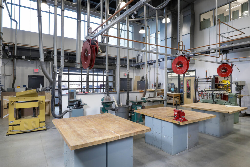 A woodworking shop with three wooden workbenches, stationary power tools, and overhead dust collection hoses with red hose reels.