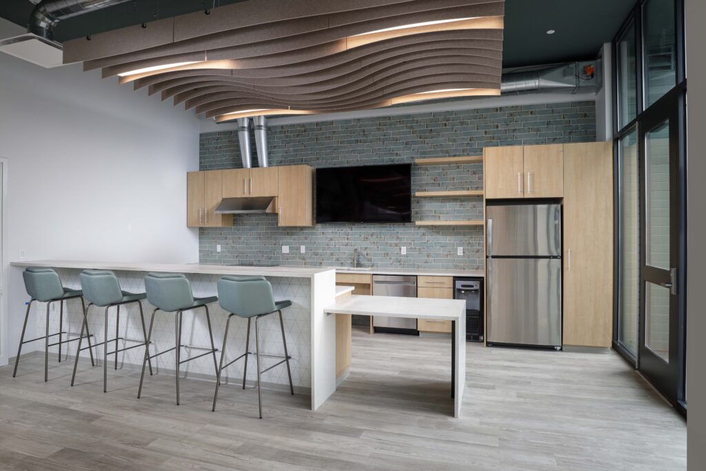Modern office kitchenette with a breakfast bar, four stools, light wood cabinets, stainless steel appliances, and a wavy acoustic ceiling feature.