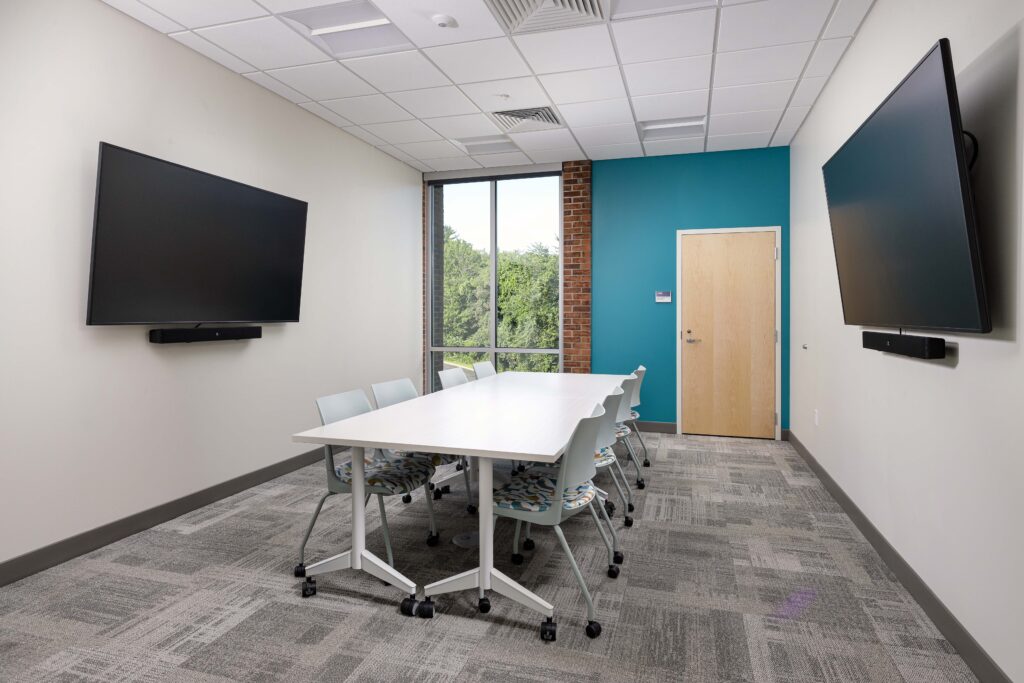 A modern conference room with a long white table, patterned chairs, and two large wall-mounted monitors.