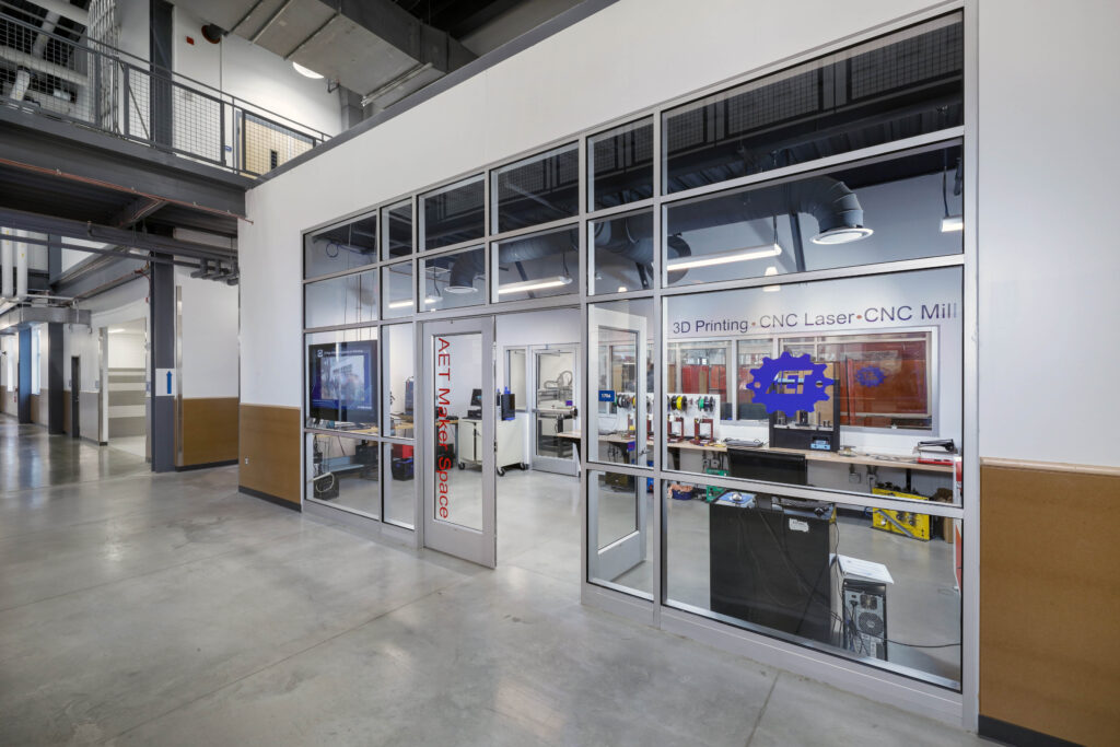 A workshop labeled “AET Maker Space” viewed through a glass-walled entrance, featuring 3D printers, CNC machines, and laser cutters.