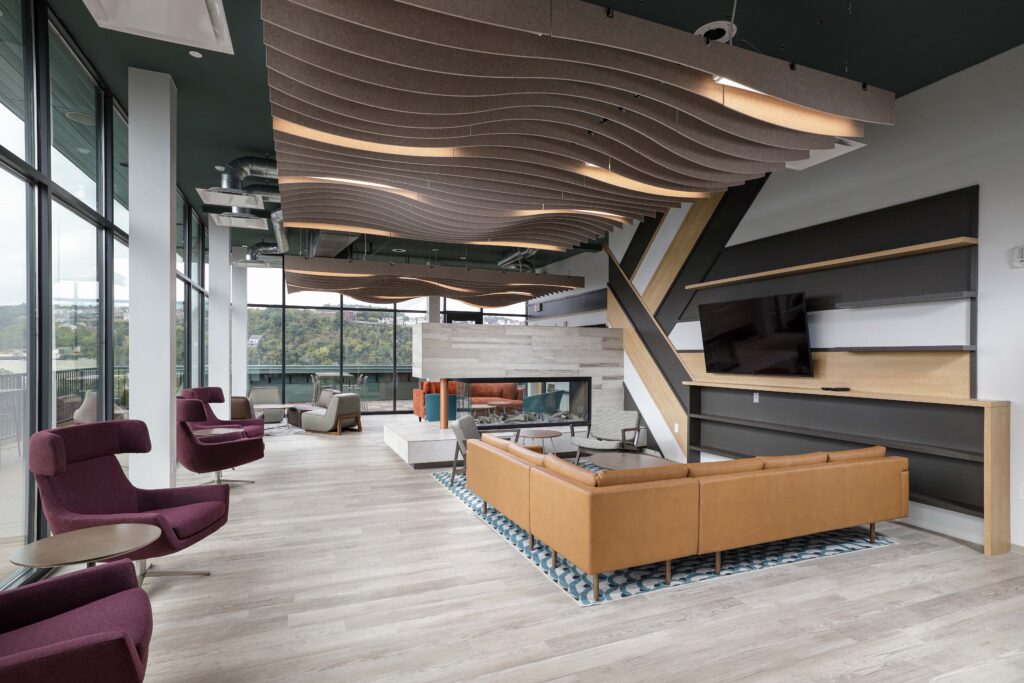 A lounge area with a tan sectional sofa, a fireplace, and several accent chairs under a wavy, suspended ceiling feature.